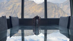 A room with a glass table reflecting the mountains and sky. There are two chairs with patterned cushions and a decorative item on the table. - J M S Guest House (Joshimath)