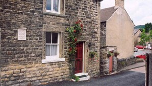 A stone building with a red door, a window with a white frame, and a sign on the wall. - Traditional 2 Bedroom Cottage in Eyam, Hope Valley (Hope Valley)