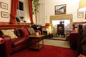 A cozy living room with a fireplace, red leather sofa, and a bookshelf. - Traditional 2 Bedroom Cottage in Eyam, Hope Valley (Hope Valley)