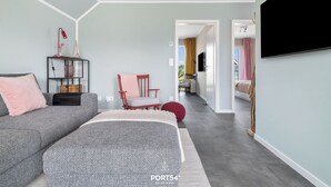 Una sala moderna con un sofá gris, un sillón rojo, un televisor montado en la pared y un vano que da a otra habitación.