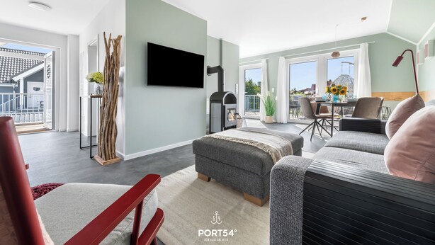 Un moderno salón con un sofá gris, un sillón rojo, un televisor de pantalla plana montado en la pared y un gran ventanal con vistas al exterior.
