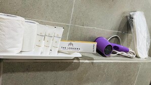 Estante de baño con artículos de aseo, incluyendo un secador de pelo morado, un kit dental y productos de ducha, loción corporal y champú de la marca The Lodgers.