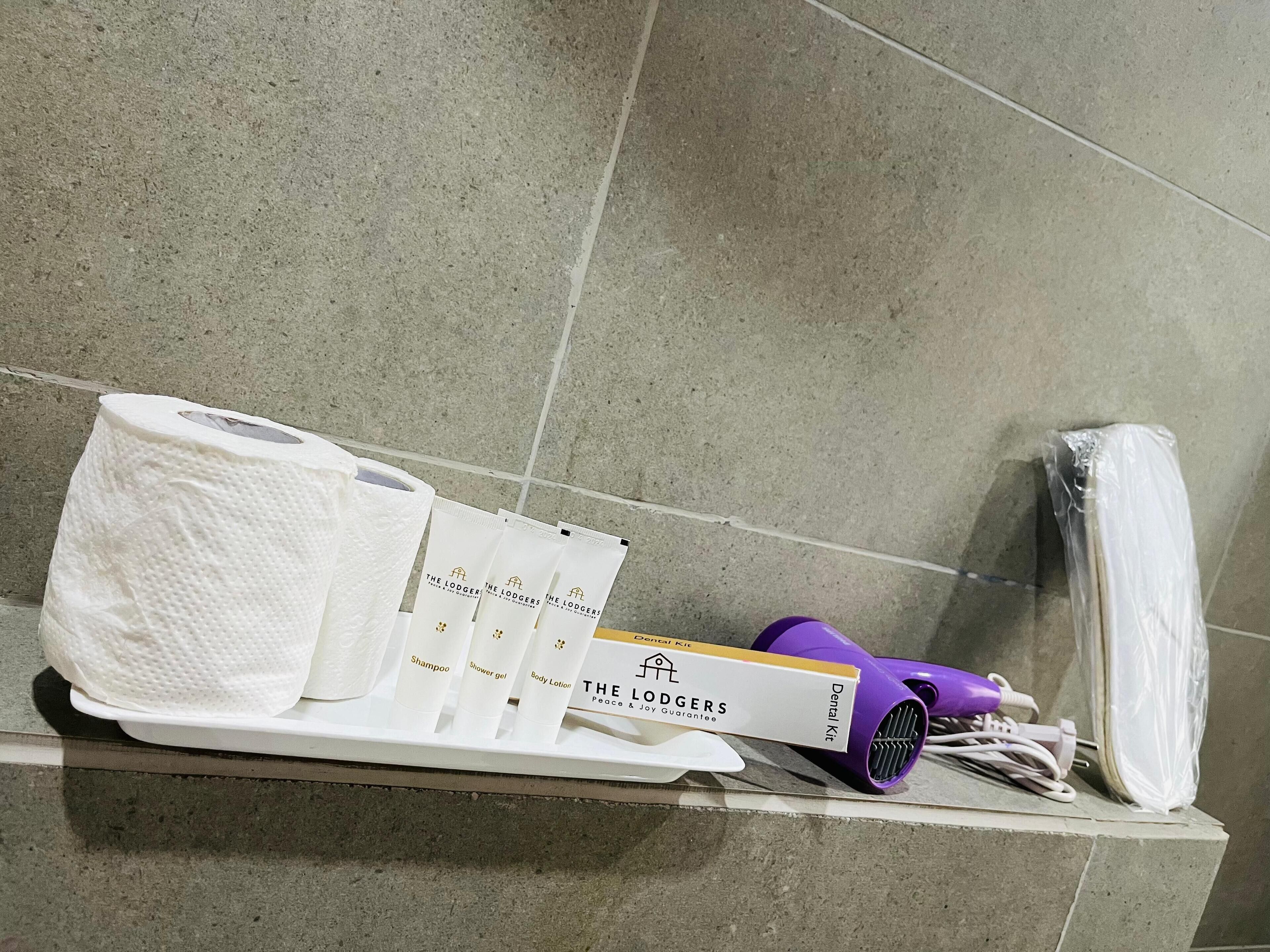 A bathroom shelf with toiletries including a pack of toilet paper, a toothbrush, a razor, and a purple hairdryer.