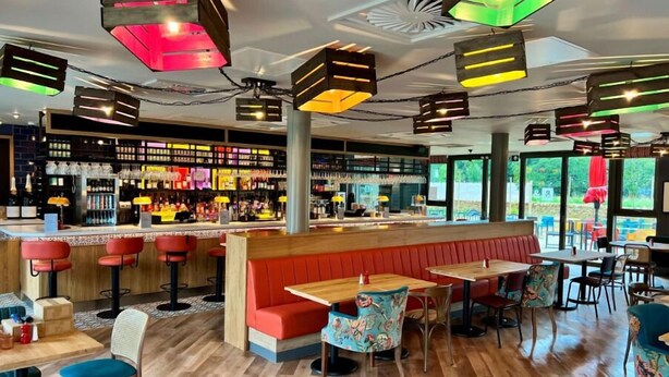 Un espace bar doté d’un comptoir en bois, de sièges rouges et de lampes colorées suspendues au plafond.