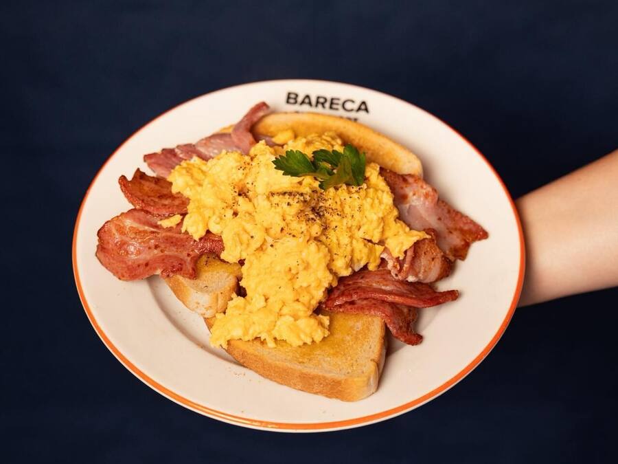 A plate of scrambled eggs, bacon, and toast with a garnish, labeled "BARECA".