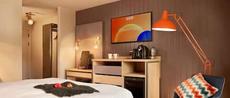 Habitación de hotel con cama, escritorio, silla, cafetera y televisor en la pared.