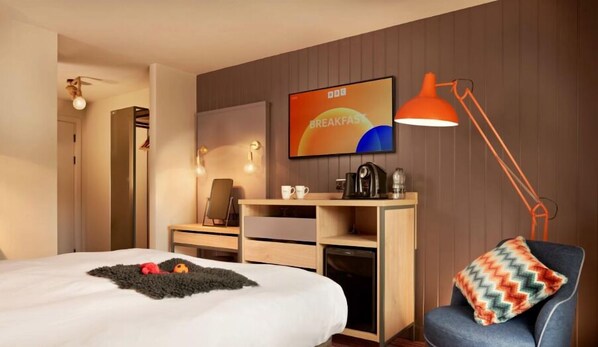A hotel room with a bed, a desk with a chair, a coffee maker, and a TV on the wall.