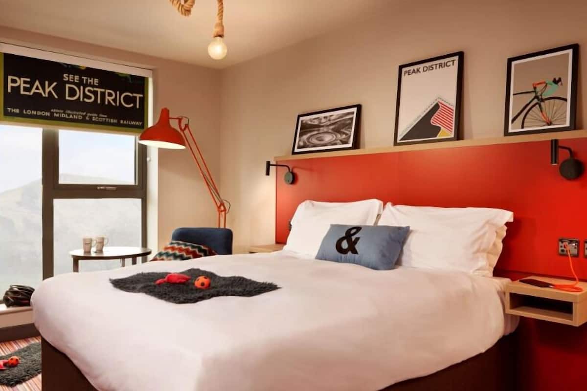 A hotel room with a large bed, a red accent wall, a window with a view of the London midland & Scottish railway, and a sign that reads "SEE THE PEAK DISTRICT".