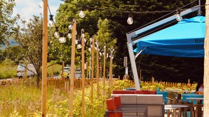 Un espace extérieur aménagé avec des tables et des chaises, entouré de verdure et surmonté d’un auvent bleu.