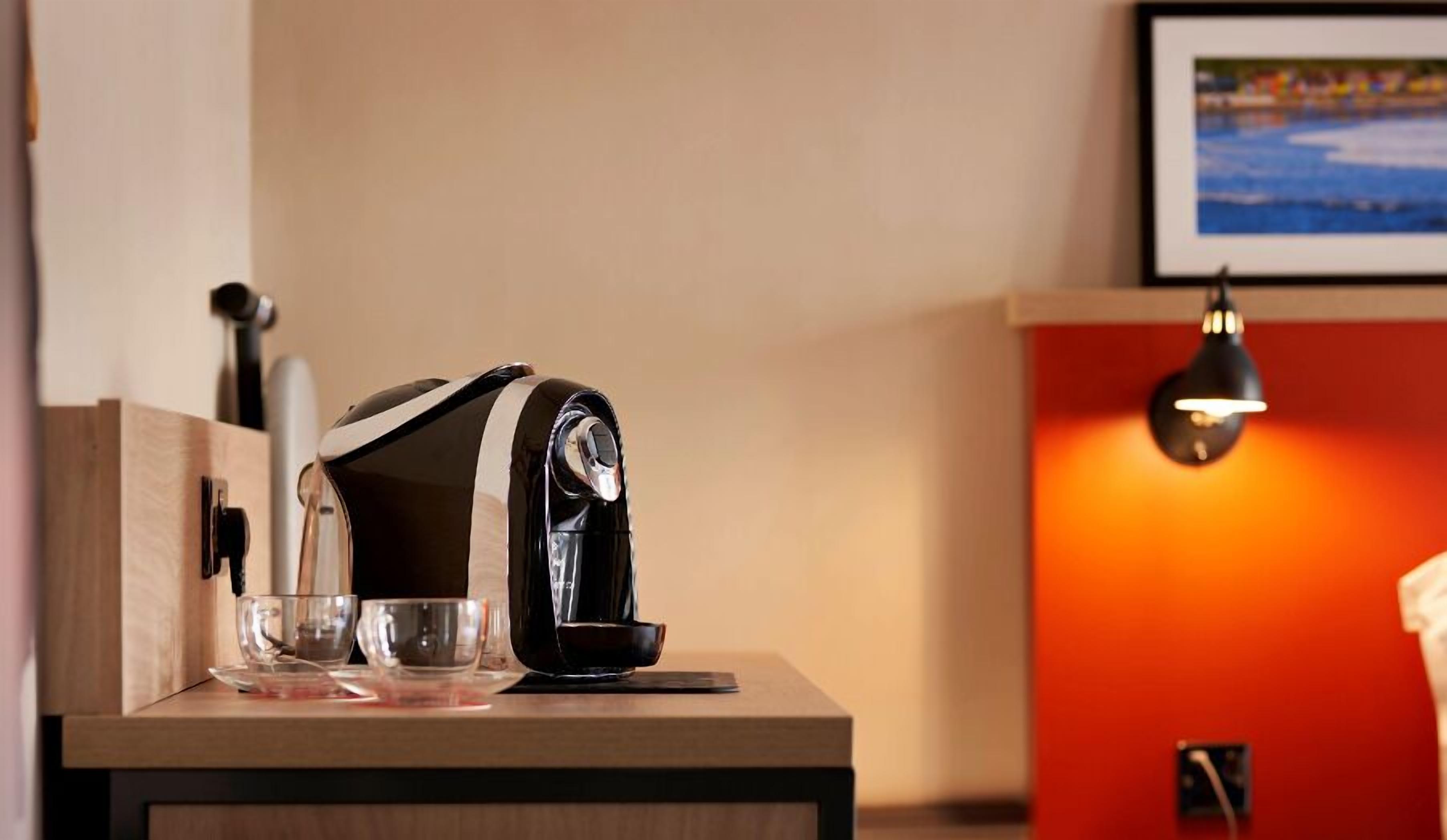 A coffee machine on a wooden counter with a glass and a cup, a framed picture on the wall, and a red wall with a lamp.