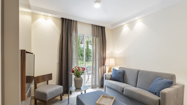 A modern hotel room with a grey sofa, a small table with a vase of flowers, a chair, and a large window with curtains. - Suite Odyssée 2 - Five Star Holiday House (Saint-Jean-Cap-Ferrat)
