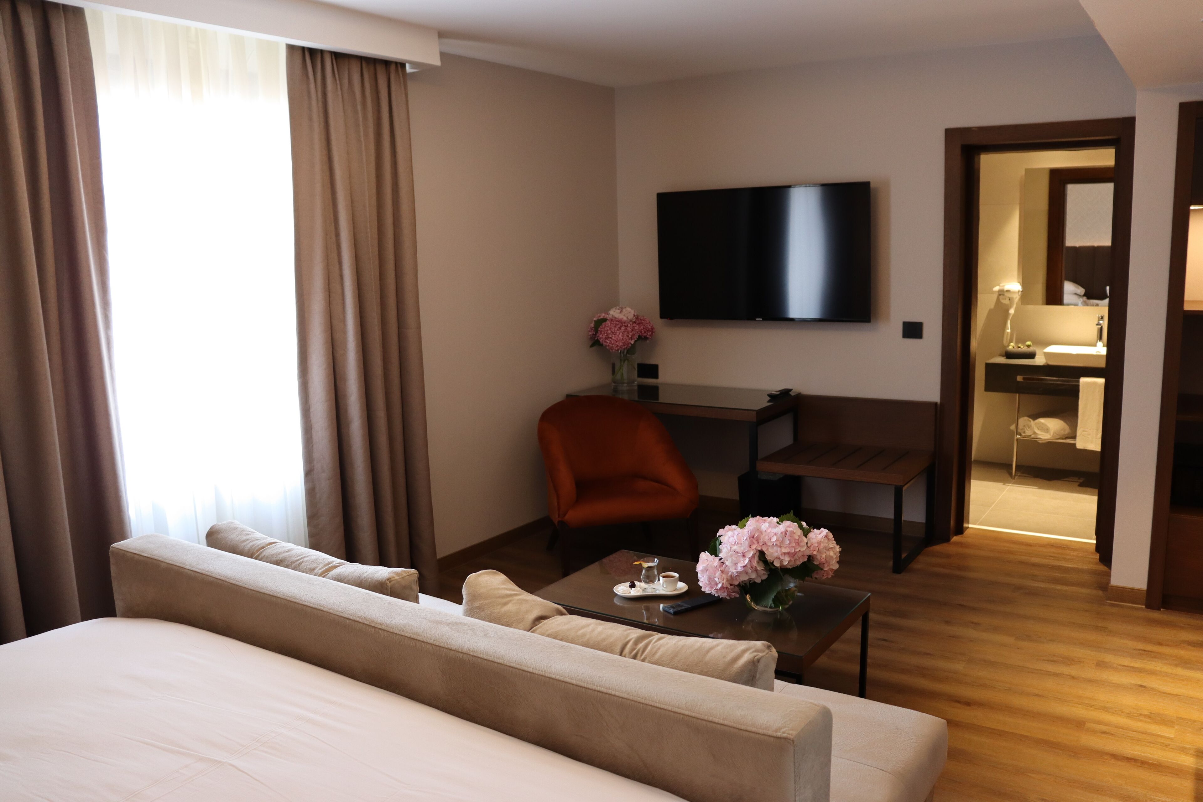 A hotel room with a large bed, a sofa, a desk with a chair, a TV mounted on the wall, and a bathroom visible through an open door.