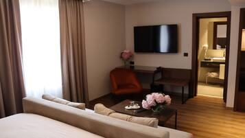 A hotel room with a large bed, a sofa, a desk with a chair, a TV mounted on the wall, and a bathroom visible through an open door.