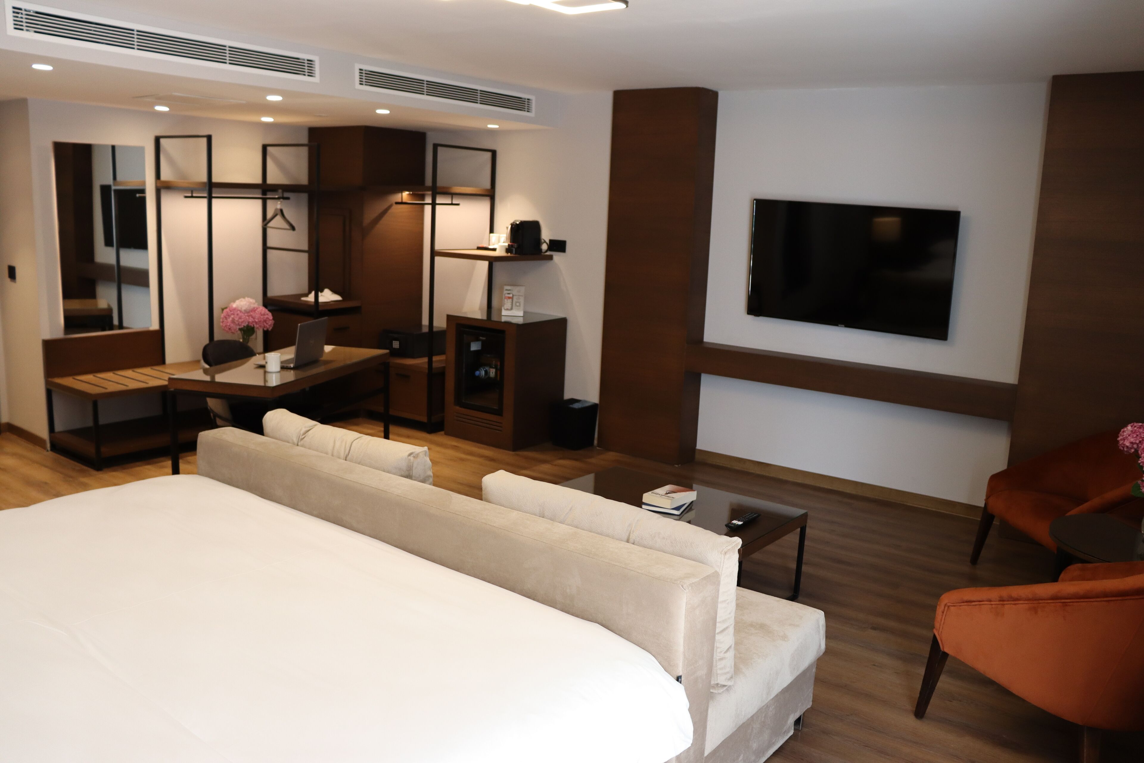 A modern hotel room with a large bed, a desk with a laptop, a TV mounted on the wall, and a seating area with a sofa and armchairs.