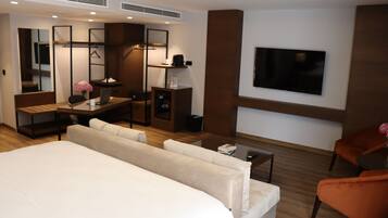 A modern hotel room with a large bed, a desk with a laptop, a TV mounted on the wall, and a seating area with a sofa and armchairs.