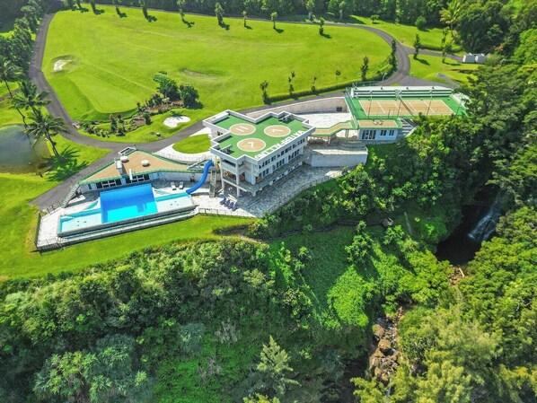 An aerial view of a resort with a swimming pool, tennis courts, and lush greenery. - Waterfalling Estate is an oceanfront compound on the Big Island unlike any other (Ninole)