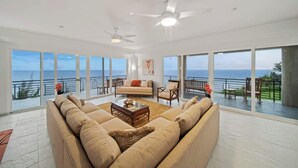 A spacious living room with a large sofa, a coffee table, and a ceiling fan. The room has large windows offering a view of the ocean. - Waterfalling Estate is an oceanfront compound on the Big Island unlike any other (Ninole)