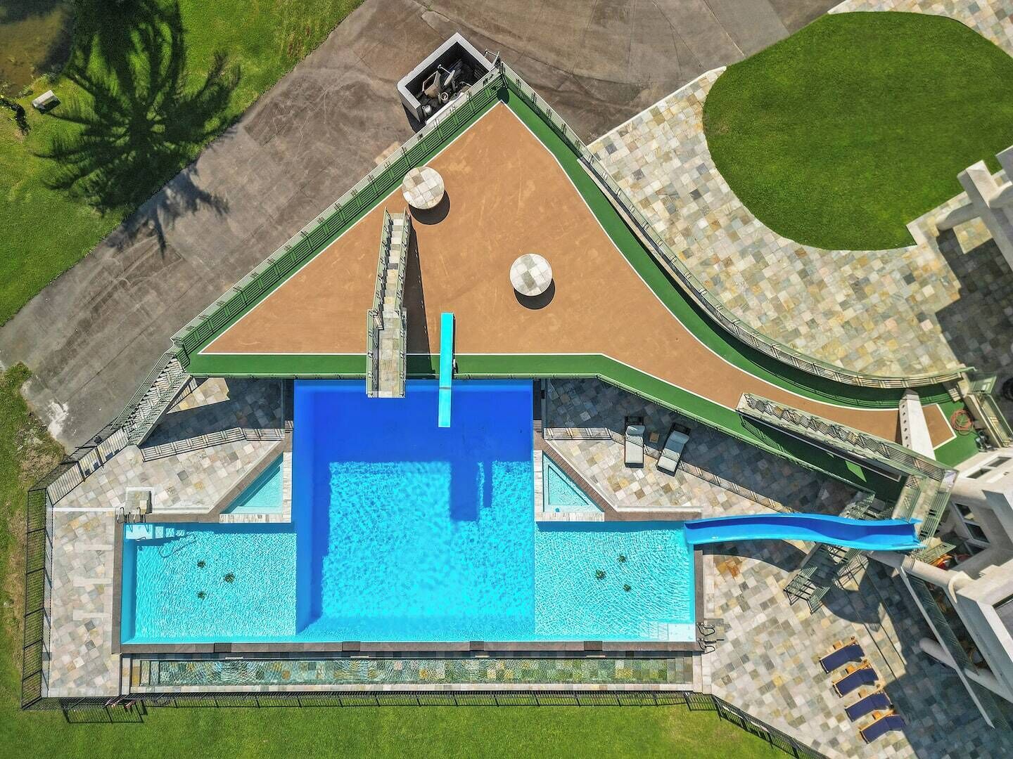 An aerial view of a resort with a large swimming pool, a tennis court, and a landscaped area with a fountain.