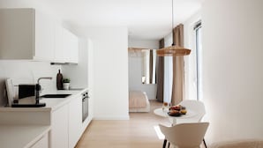 A modern kitchen with white cabinetry, a dining area with a round table and chairs, and a living space with a sofa and a large window. - BOHO City Suites Augarten (Vienna)