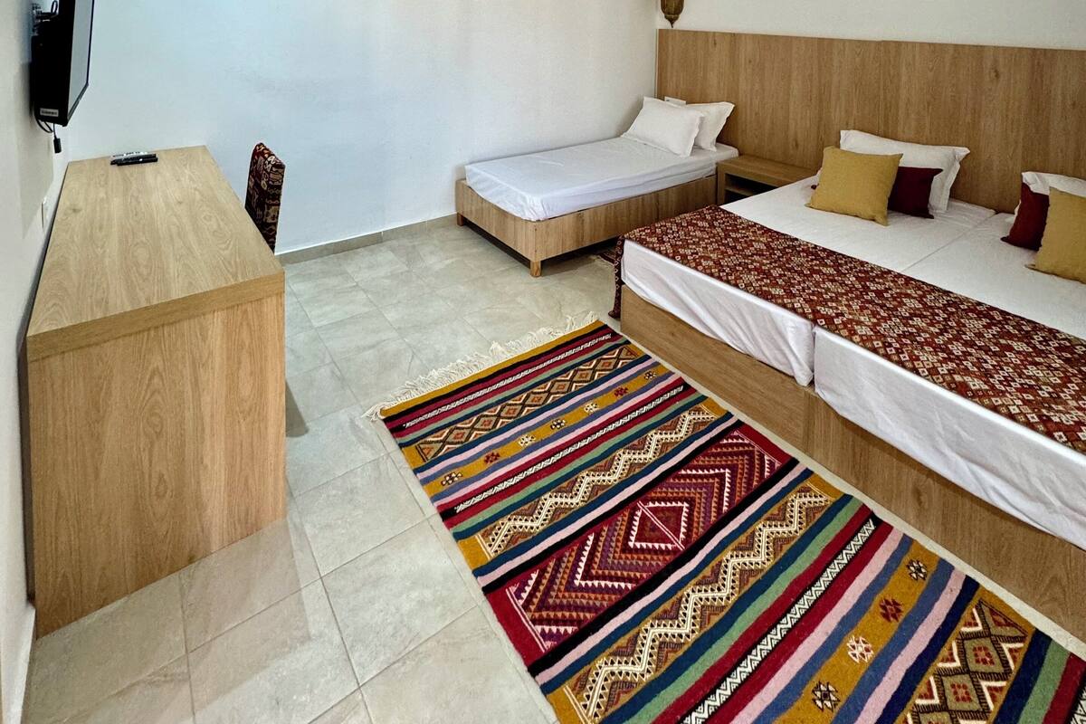 A hotel room with a wooden desk, a bed with patterned bedding, a colorful rug, and a wall-mounted TV.