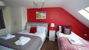 A bedroom with a red accent wall, a chandelier, a bed with white bedding, a bedside table with a lamp, and a small white drawer unit. - 59B Comfortable Home Sleeps 8 (Hounslow)