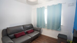 A grey leather sofa with two red cushions, a window with light blue curtains, and a small wicker basket. - 59B Comfortable Home Sleeps 8 (Hounslow)
