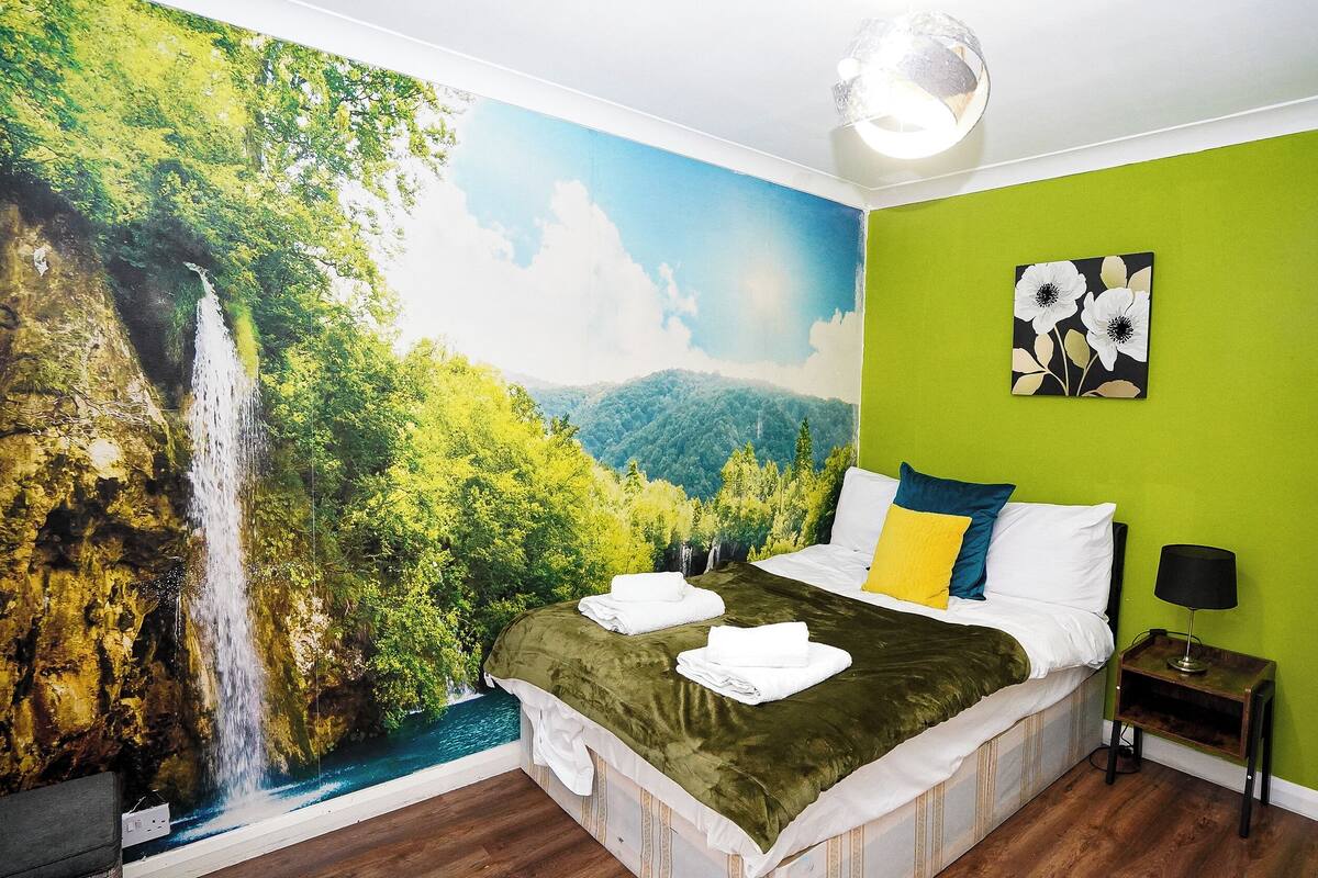 A bedroom with a mural of a waterfall, a bed with a green bedspread, white and yellow pillows, and a nightstand with a lamp.