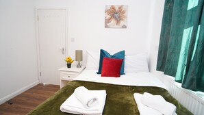 Una habitación de hotel con cama, mesita de noche con lámpara, jarrón con flores y ventana con cortinas.