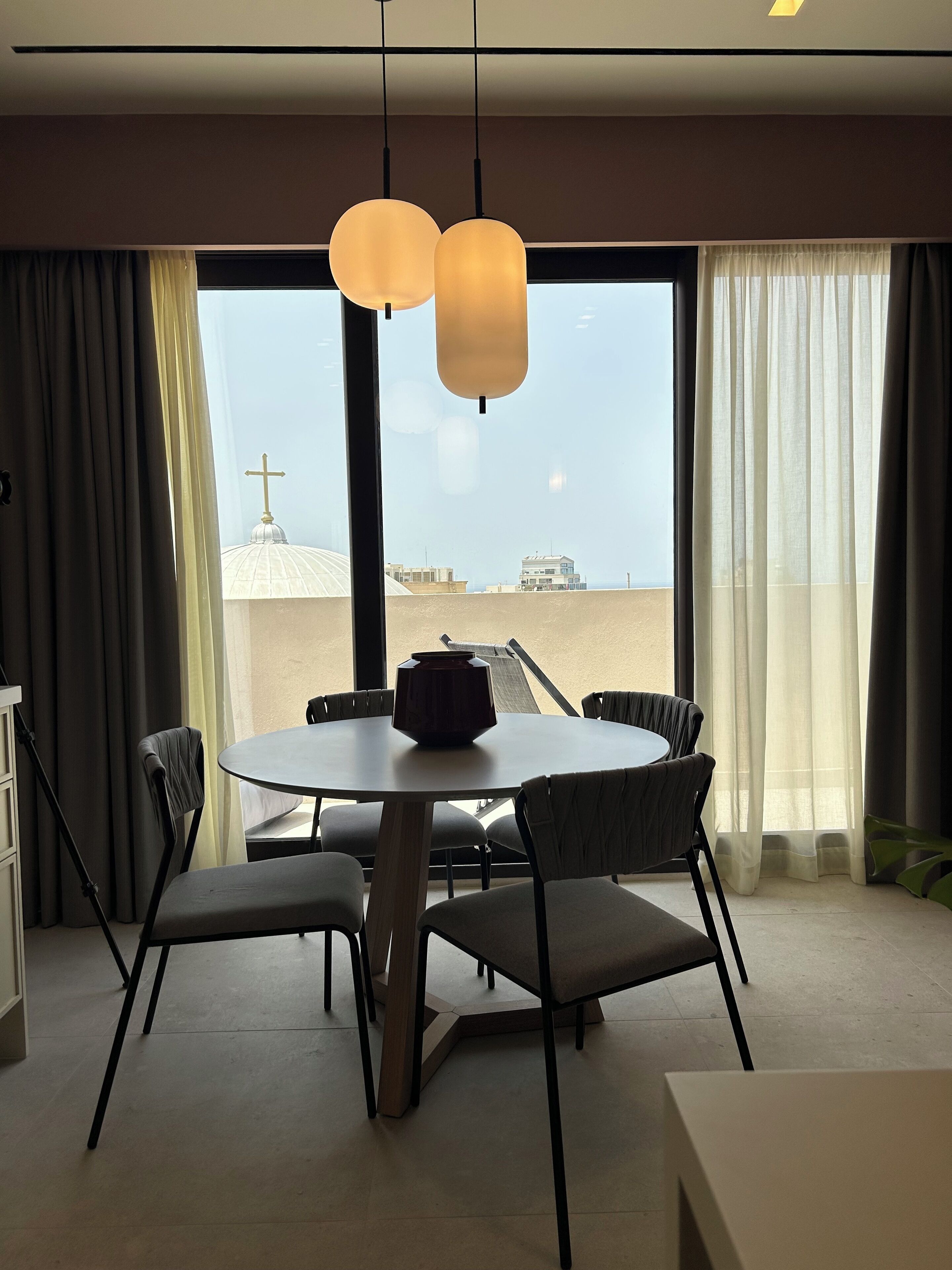 Ein moderner Essbereich mit einem runden Tisch, sechs Stühlen und Pendelleuchten. Ein großes Fenster bietet Blick auf ein Gebäude mit einer Kuppel und einem Kreuz.