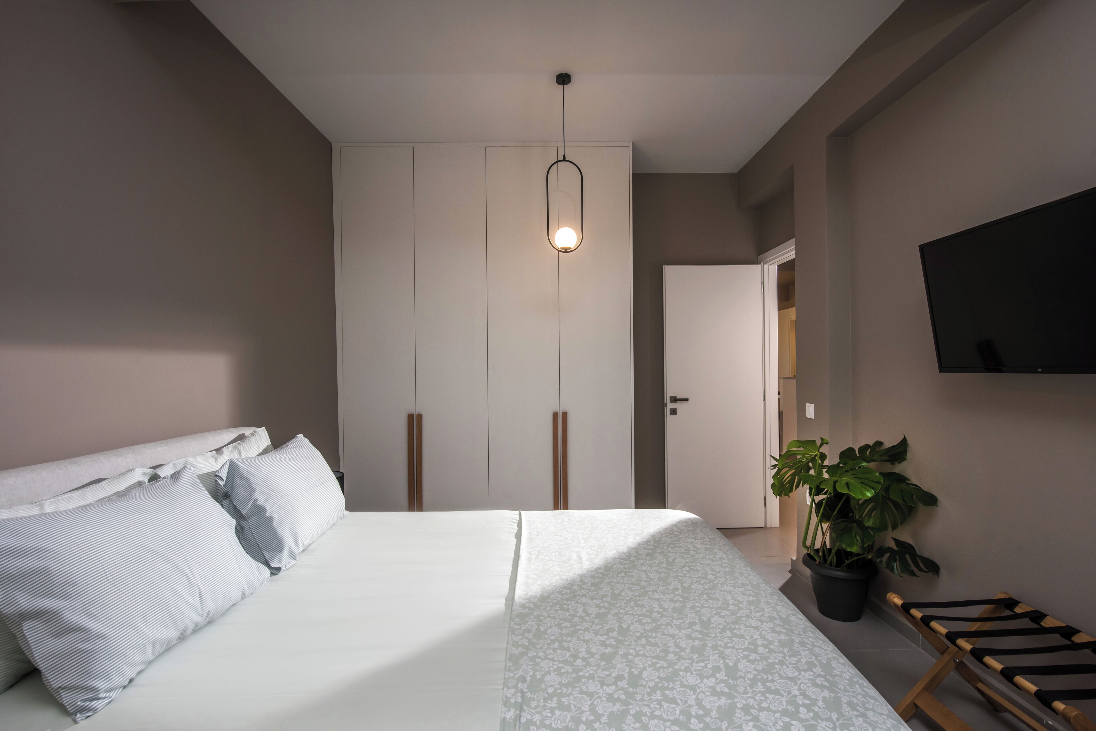 A bedroom with a bed, bedside table, potted plant, and a TV mounted on the wall.