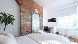 A bedroom with a brick wall, a large mirror, a bed with white bedding, a desk with a chair, and motivational wall art. - Close to Tube - Free Parking - Pet Friendly (London)