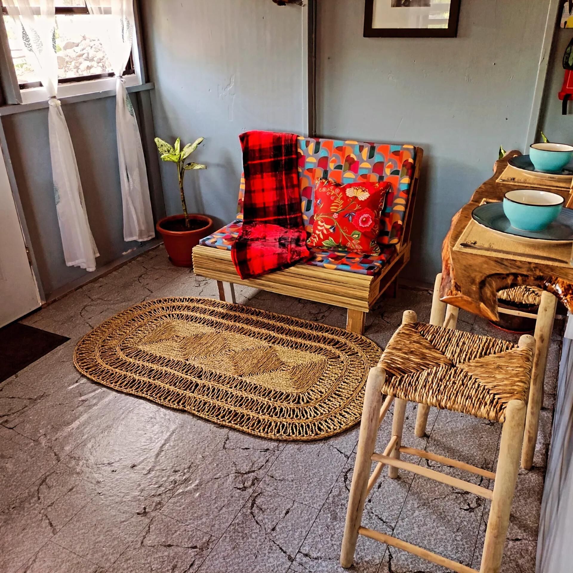 Una habitación con un tapiz tejido, una silla de madera, un sofá con una manta roja, una planta en maceta y una mesa con cuencos.