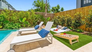 Un patio trasero con piscina, dos sillones de jardín con cojines, una pequeña mesa con bebidas y una manta a cuadros sobre el césped.