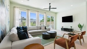 Uma sala de estar moderna com um sofá, mesa de centro e cadeiras, um ventilador de teto e uma TV na parede.