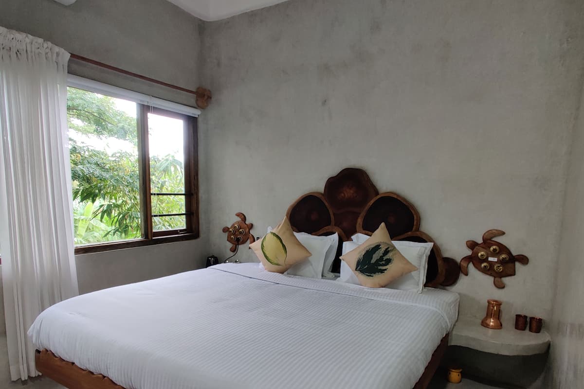 Quarto com cama de madeira, roupa de cama branca, janela com cortinas e mesa de cabeceira com objetos decorativos.