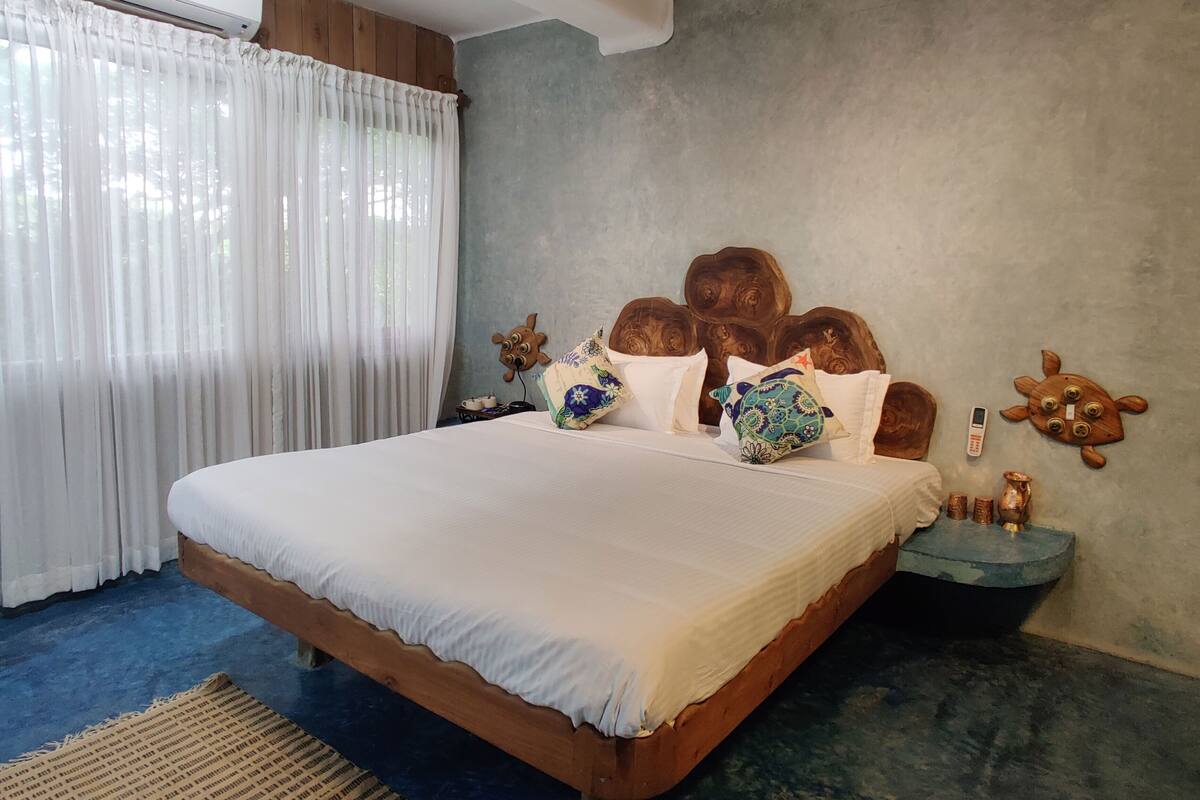 Um quarto com uma cama grande, um ventilador de teto, um ar condicionado, uma cabeceira de madeira, arte decorativa na parede e uma mesinha de cabeceira com um telefone e um vaso.