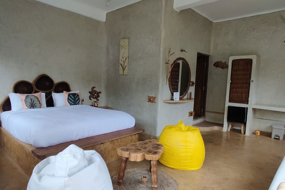 Quarto com uma cama, ventoinha no teto, um banco de madeira, um pufe amarelo e uma mesa pequena.