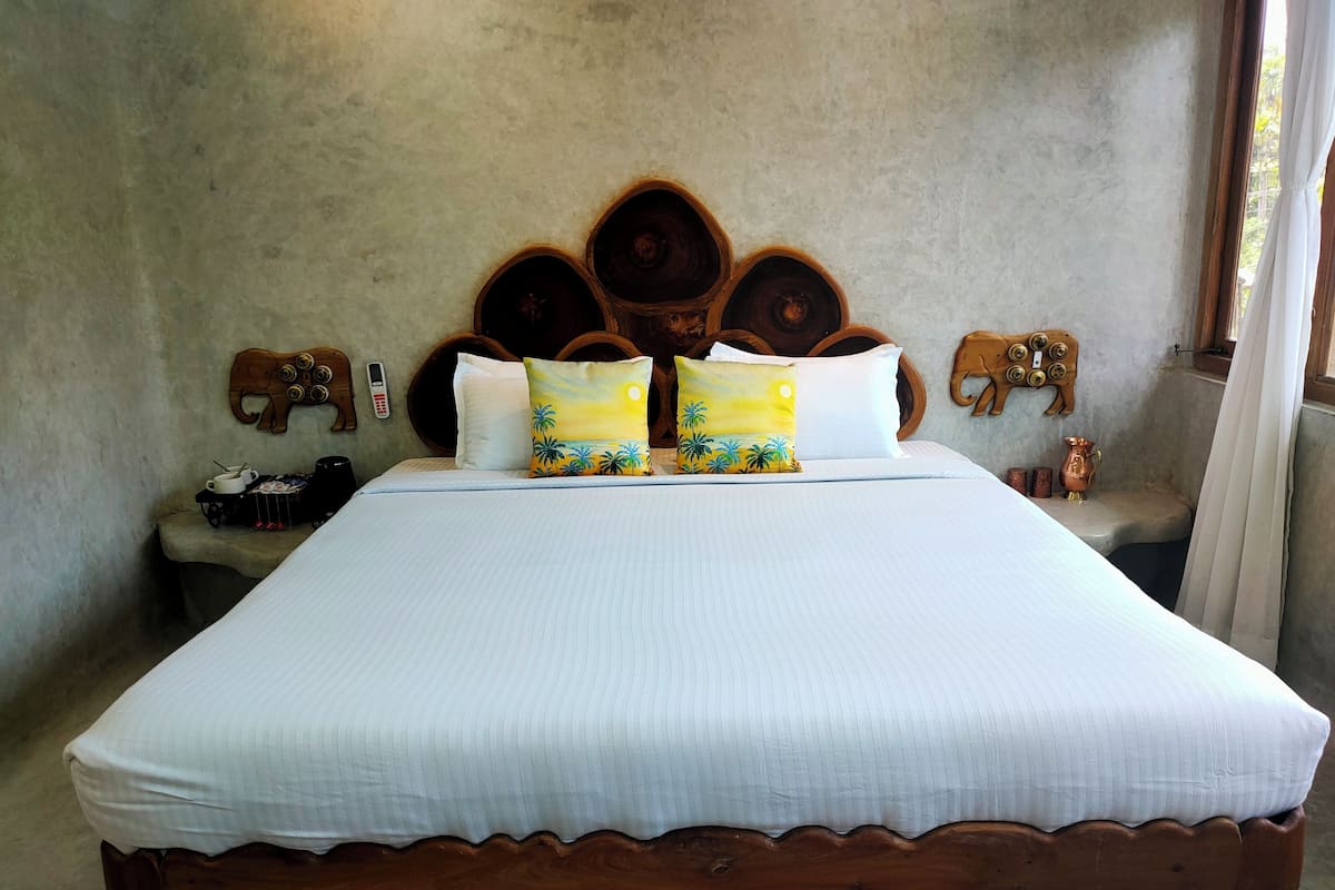 Uma cama grande com cabeceira de madeira, duas almofadas, mesa de cabeceira com um telefone e um abajur, e uma pequena estátua no criado-mudo.
