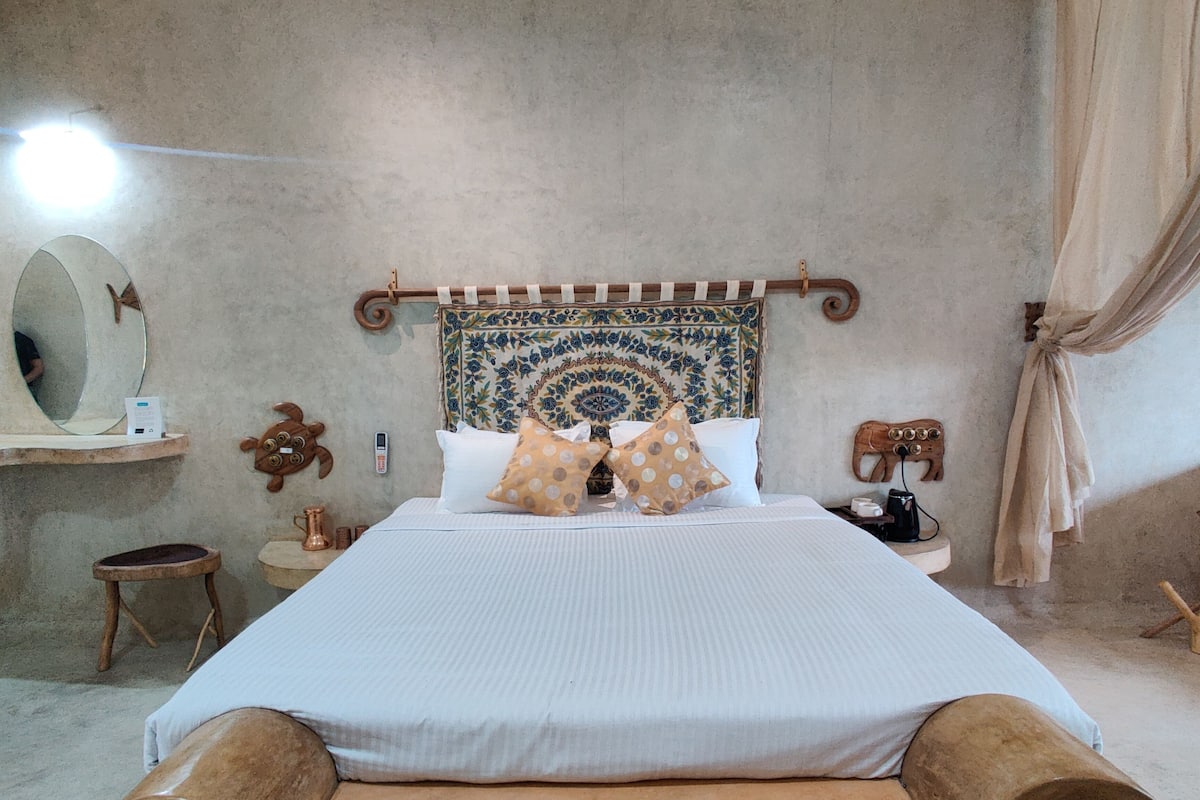 Uma cama com cabeceira de madeira, uma mesinha de cabeceira, um espelho e uma cortina.