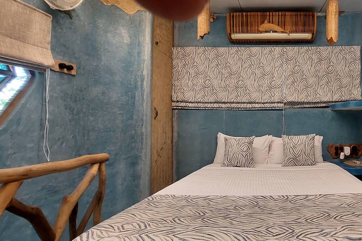 Quarto com cama, cadeira de madeira, cabeceira estampada e pequena mesa com espelho.