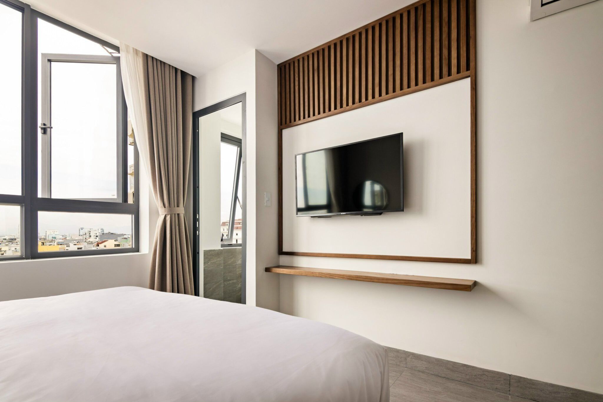A modern hotel room with a large bed, a TV mounted on the wall, and a city view through the window.