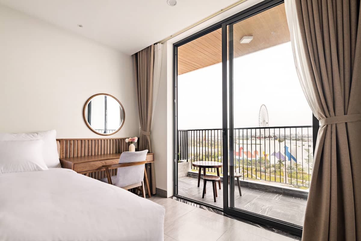 Une chambre d’hôtel avec un lit, un bureau en bois, un miroir rond et un balcon offrant une vue sur la ville.