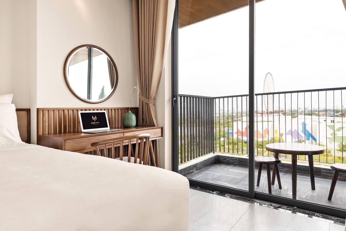Une chambre d’hôtel comprenant un lit, un bureau avec un ordinateur portable, un miroir rond et un balcon avec une table et des chaises.