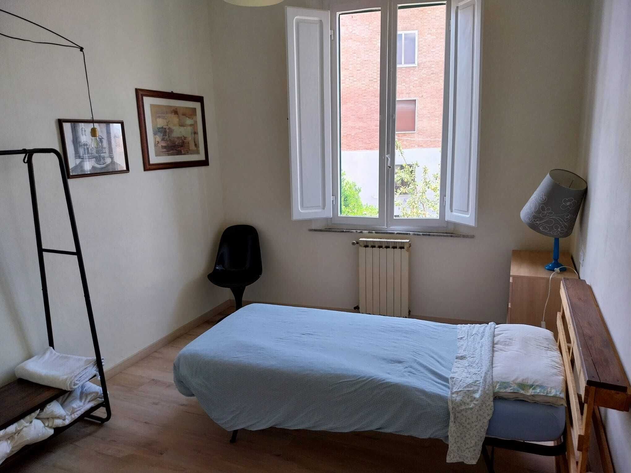 A bedroom with a single bed, a radiator, a chair, a desk with a lamp, and two framed pictures on the wall.