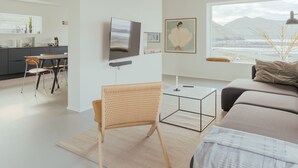 A modern, minimalist living room with a large bed, a dining area, and a kitchen in the background.