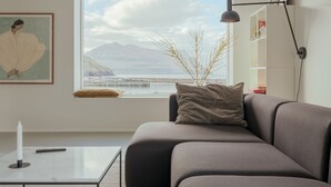 A modern living room with a large window, a grey sofa, a small table, and a lamp.