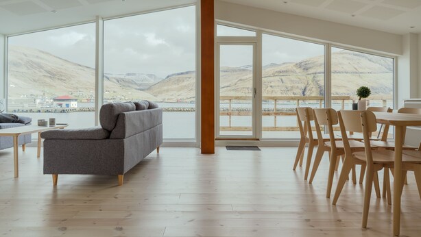 A modern interior with large windows, a grey sofa, wooden flooring, and a dining area with wooden chairs.