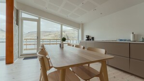 A modern kitchen with a large island, wooden dining table, and chairs. There is a view of a mountainous landscape through large windows.