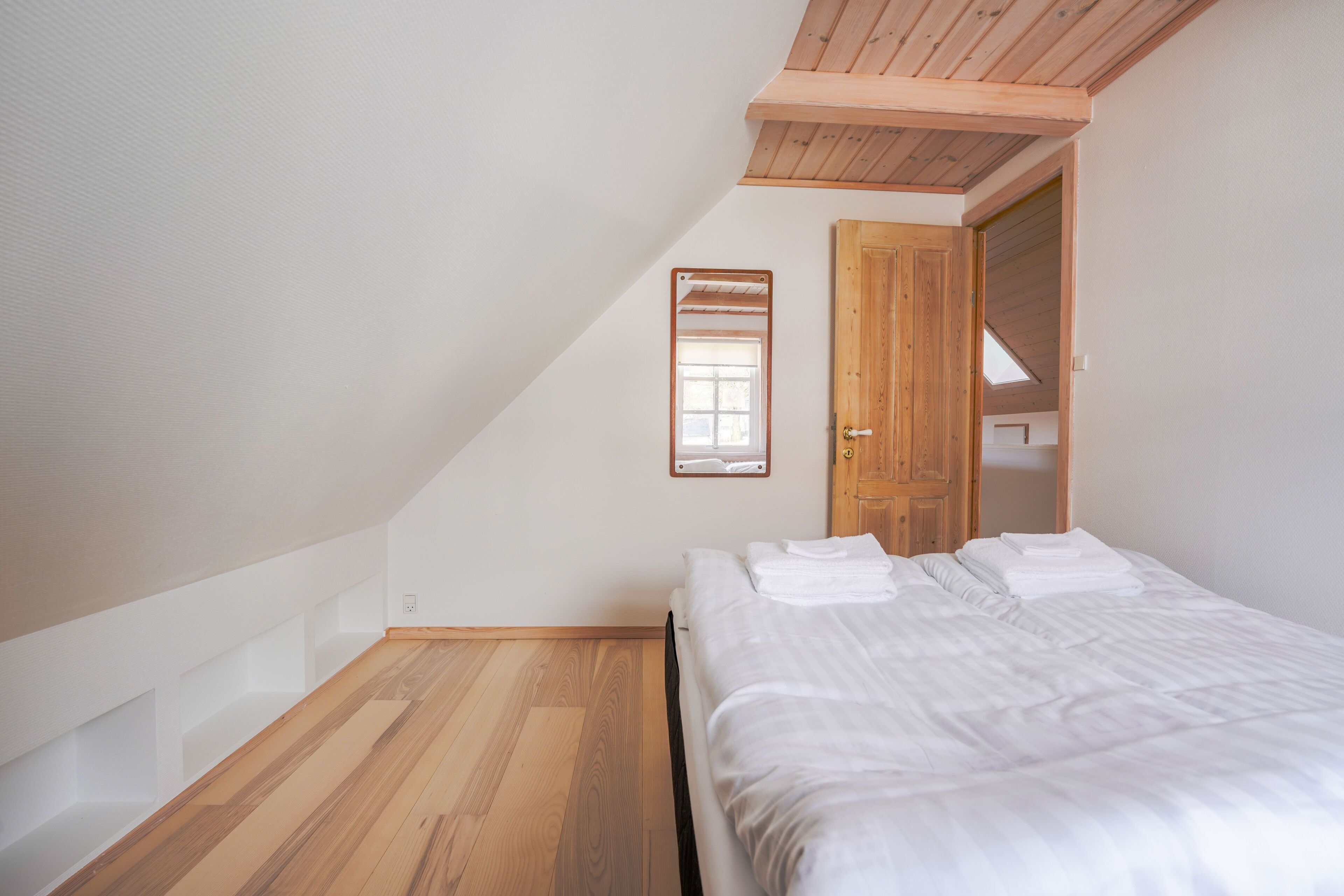 A bedroom with a wooden floor, a bed with white linens, a wooden door, and a window.