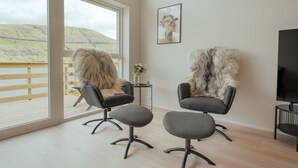A modern room with large windows, two chairs with fluffy fur throws, a small table with a vase, and a framed picture on the wall. - Luxurious Waterfront Retreat | Spectacular Views (Vestmanna)